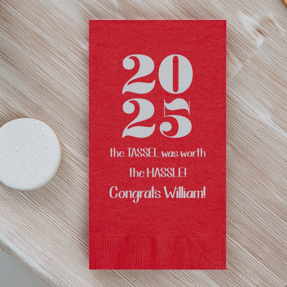 A red color guest towel napkin personalized with bold block numbers "2025" for a graduation celebration in silver print. Three lines of custom text include the wording "The Tassel was worth the Hassle!" and the graduate's name. The guest towel is laying an a bamboo textured sink top for inspiration on what to have in bathrooms for graduation.