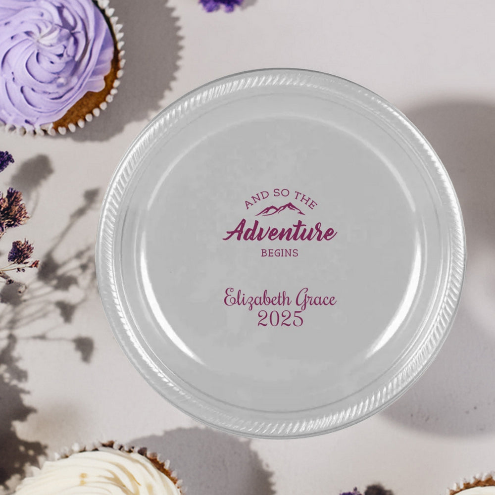 A silver plastic plate custom printed for a 2025 graduation party in maroon print setting on a table next to cupcakes and floral accents. The plate is personalized with a mountain top design with the working "And so the adventure begins" building excitement for the graduate's future plans. Below the design, printed in a fun script font is the graduate's name and graduation year "2025"