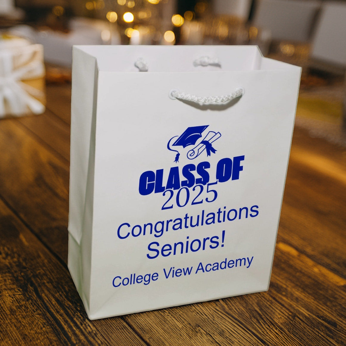 The white Euro Tote gift bag with a white braided handle in this image was personalized in blue print for graduating student gifts and is setting on a dark wood table next to a blue and a gold graduation cap. The design on the gift bag features a graduation cap above large bold italic wording "Class of 2025" with three lines of custom text below that read "Congratulations Seniors!, College View Academy.