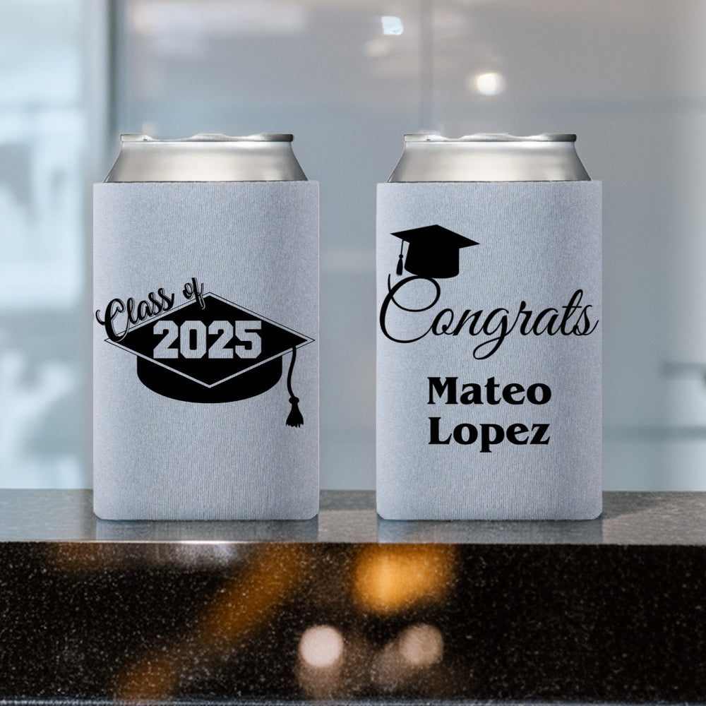 Two silver frost color can coolers personalized for a graduation celebration setting on a dark quartz countertop. The can cooler sleeve on the left is customized with a grad cap design featuring "Class of" text in script font on the upper left corner of the cap and "2025" in the center of the cap. The can sleeve on the left in customized with a "Congrats" calligraphy design with a graduation cap over the letter "C". The graduate's name is printed below the design in a bold modern italic font.