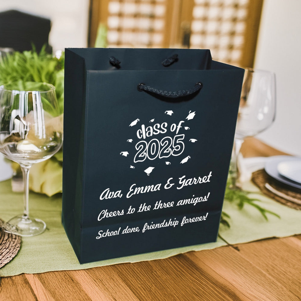 A blue Euro Tote gift bag custom printed for a graduation party in white print is setting on a graduation dinner table decorated with light green tablecloth and a green floral centerpiece in the background. The gift bag is personalized with a design featuring the wording "class of 2025" surrounded by flying graduation caps. Below the design are three lines of text including the names of three graduates and the phrase "Cheers to the three amigos! School done, friendship forever!"
