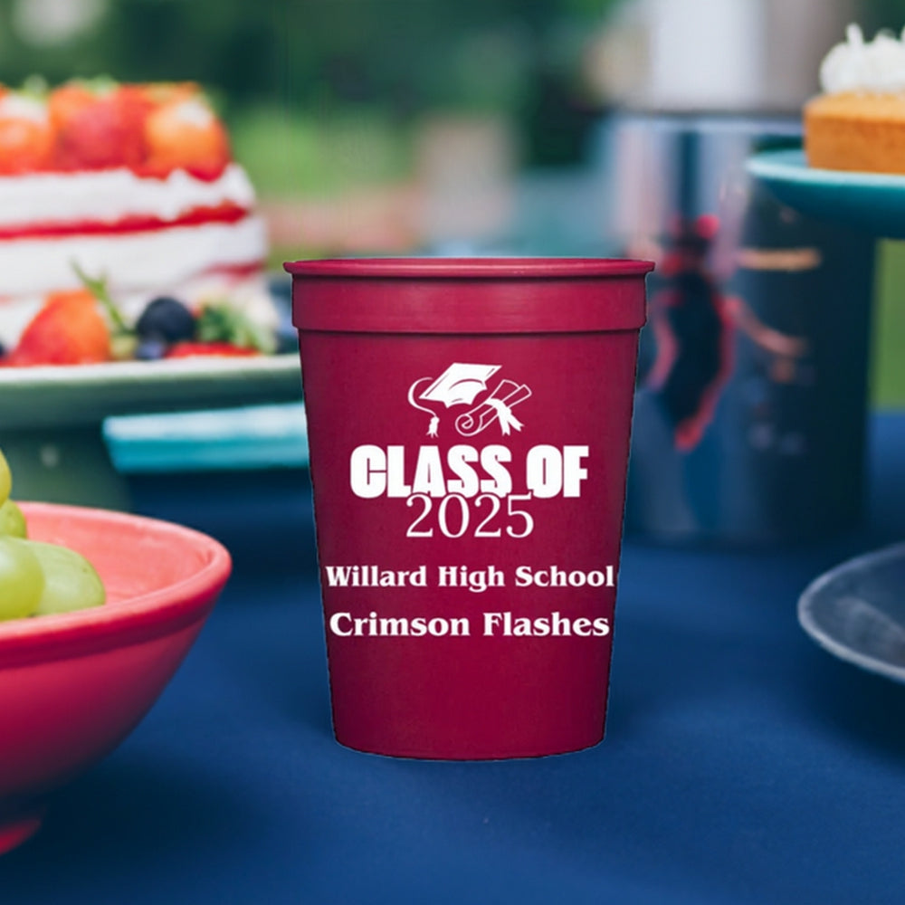 A small red plastic cup designed for a graduation celebration is customized with a design: "Class of 2025" in bold white text, along with a graduation cap and diploma graphic. The graduates' high school name and mascot name are printed below the design in "Sociable" font. The cup is set on a blue tablecloth, surrounded by party food, including a strawberry-topped cake and a bowl of grapes at an outdoor seniors graduation party.