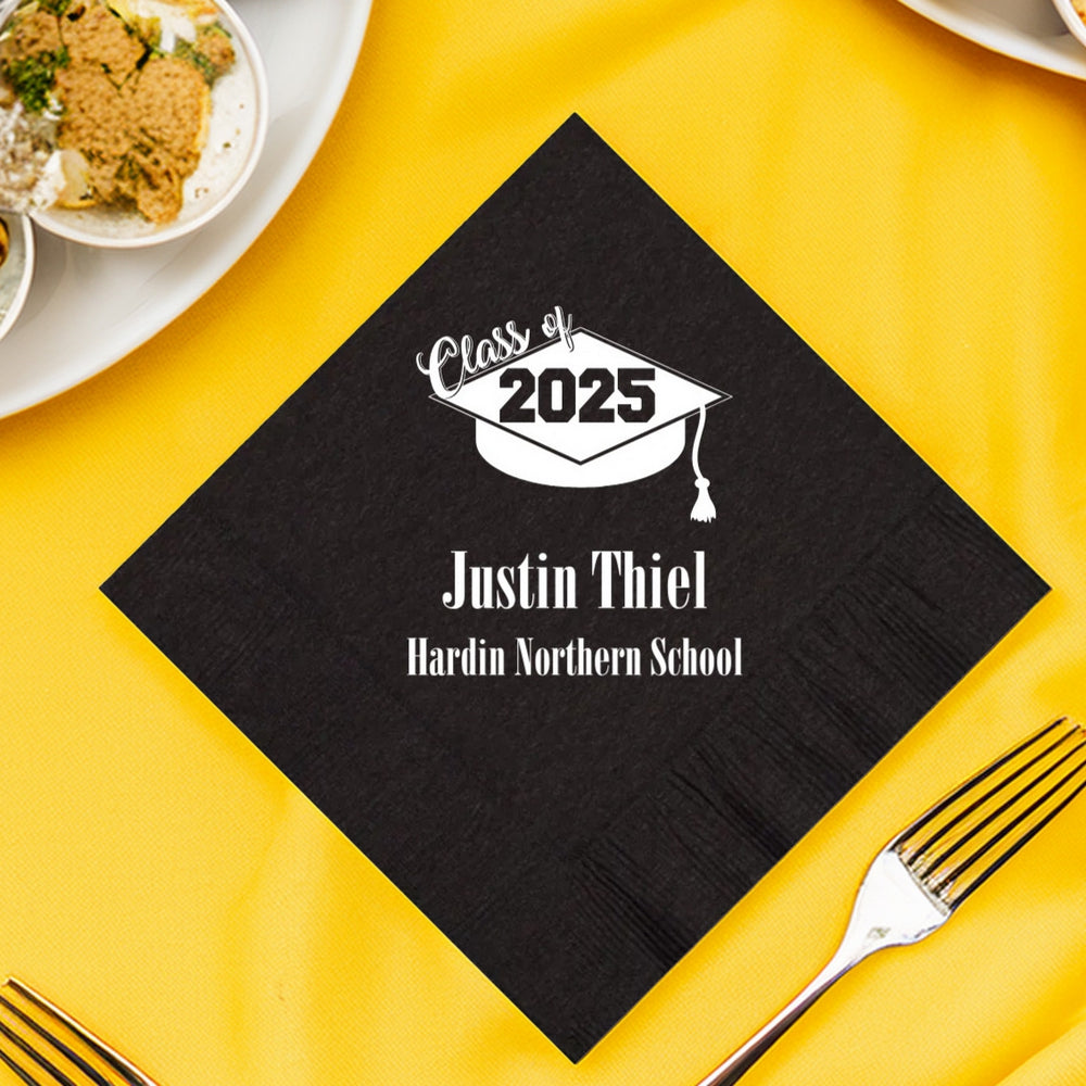 A black, diamond-shaped graduation napkin personalized in white print with a graduation cap with a tassel graphic at the top, accompanied by the text "Class of 2025" in a trendy cursive font for "Class of" and a bold, block font for "2025." Below the graduation cap, the graduate's name is prominently displayed in large, bold, white letters. Under the name, the high school name is printed in smaller, bold, white letters. The napkin is laying on a yellow tablecloth with snack foods and forks.