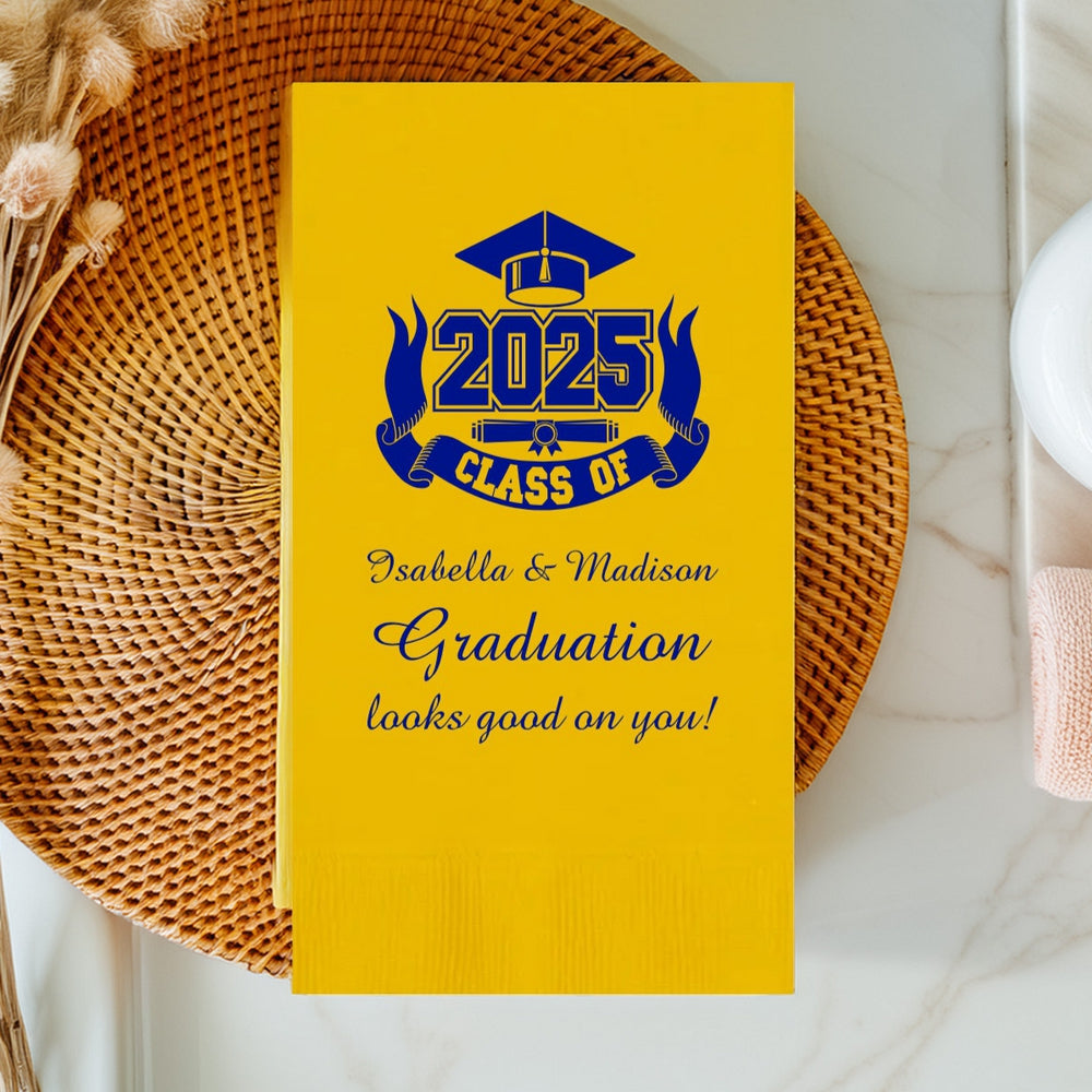 A Yellow paper hand towel napkin personalized for a graduation party is setting on a wicker place mat on a bathroom vanity top. The napkin is personalized in bright blue print with a Class of 2025 graphic that features a graduation cap above "2025." A ribbon with the wording "Class of" wraps around the sides and bottom of 2025 with a diploma centered between the ribbon and "2025." Three lines of text printed in a beautiful script font include two graduates' names and wording "Graduation looks good on you!"