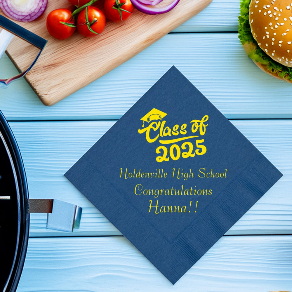 A blue, diamond-oriented luncheon napkin personalized for a graduation in yellow print. At the top is a graduation cap with a tassel graphic with the text "Class of 2025" in cursive font for "Class of" and a bold, block font for "2025," below. Below the design is three lines of custom text that includes the high school name, written in smaller serif font, the word "Congratulations", and the graduate's name in larger, bold, lettering with two exclamation points.