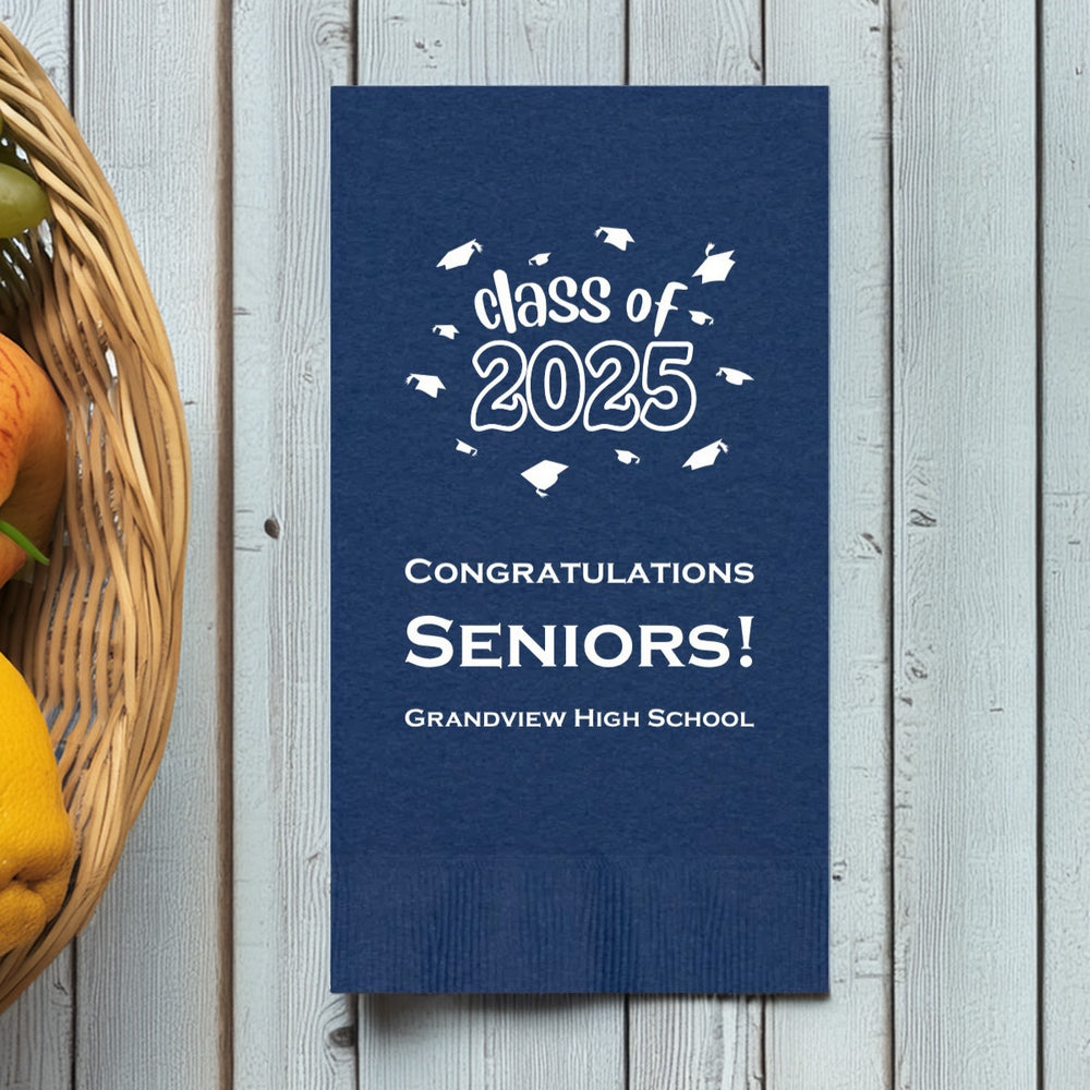 A navy blue paper guest towel napkin personalized for a graduation party is laying on a white wood table next to a basket of fruit. The guest towel is personalized in white print with a fun graphic design of the wording "class of 2025" surrounded by flying graduation caps. Below the design are three lines of custom text in white print that reads "Congratulations SENIORS!" along with the graduate's high school name "Grandview High School"