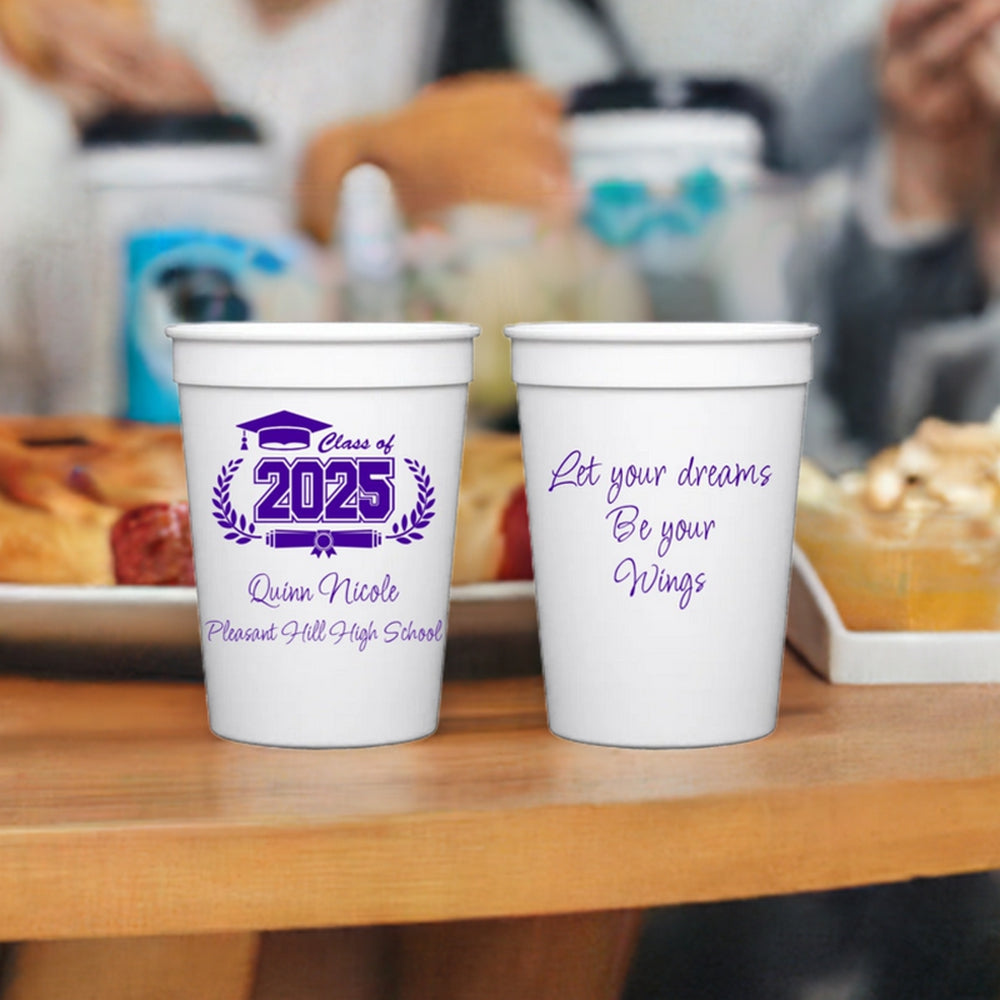 Two small white plastic cups personalized for a graduation party with purple print are sitting on a wood table in front of food and drinks. The cup on the left reads "Class of 2025" with a graduation cap graphic and laurel wreath design, along with the graduate's name and high school printed in "Flirty" font. The cup on the right is printed in "Flirty" font with a quote: "Let your dreams Be your Wings."