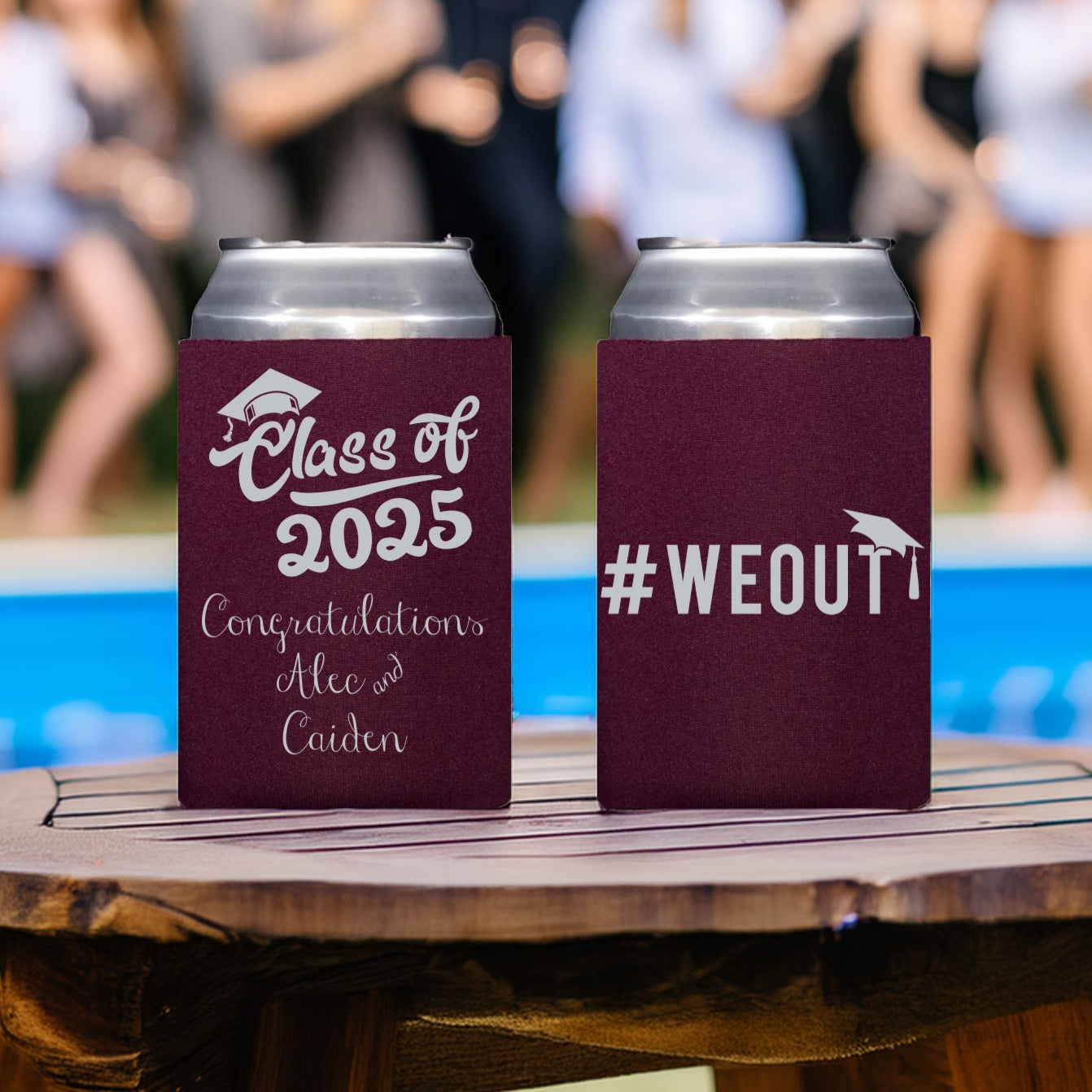 Two burgundy color graduation can sleeves are setting on a wood tabletop with a swimming pool and young people mingling in the background. The can sleeve on the left is personalized in silver print with a script "Class of 2025" design featuring a graduation cap over the "C" in "Class". A message "Congratulations" is displayed below the design with two names for a double graduation party. The can sleeve on the right is personalized with a "hashtag WEOUT" design with grad cap in silver print.