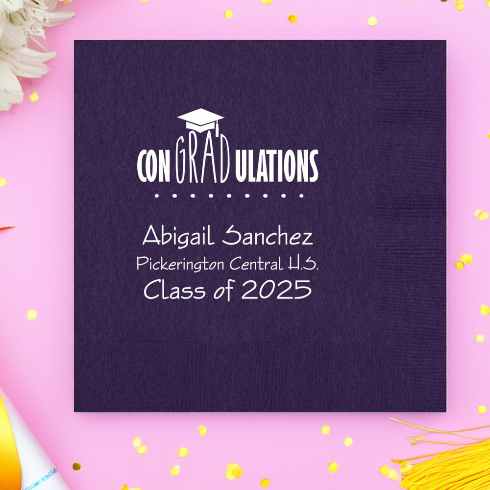 A dark purple paper dinner napkin personalized for a graduation party laying on a pink tablecloth sprinkled with gold confetti. The napkin is customized in a square orientation in white print with a "CONGRADULATIONS" graphic for a fun play on words to congratulate the high school graduate. A grad cap is positioned above "GRAD" with a series of dots underneath the design. Below the design are three lines of text in a fun italic font that display the graduate's name, high school and "Class of 2025".