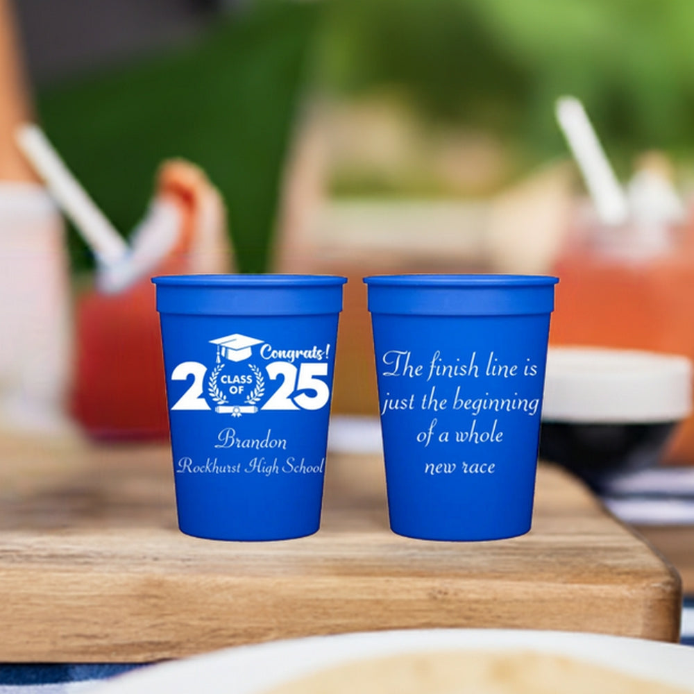 Two 12 ounce blue plastic stadium cups personalized for a 2025 grad party are setting on a cutting board on a table at an outdoor setting. The left cup is personalized in white print with bold "2025" compliments with a grad cap, diploma and wheat wheat stalks around the wording "Class Of". The graduate's name and high school are printed below the design in "Blissful" font. The right cup is personalized in white print with 4 lines of text that read "The finish line is just the beginning of a whole new race."