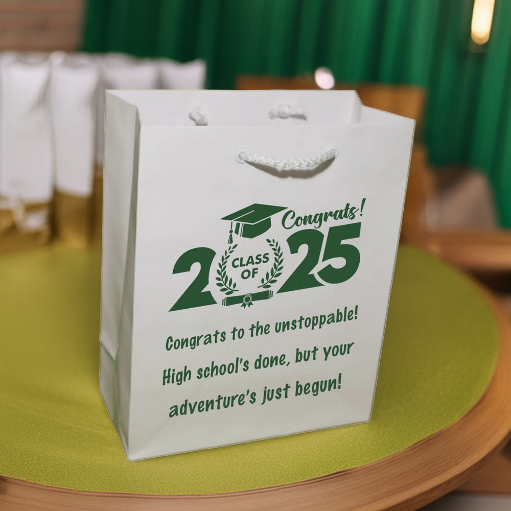 A glossy white tote bag personalized in green print for giving gifts to students after their graduation ceremony is setting in front of a row of gift bags on an a table with a green tablecloth. A bold design exclaims "Congrats! Class of 2025." The words "Class of" are surrounded by two wheat stalks with a grad cap above and a diploma below the wording. Three lines of text below the design read "Congrats to the unstoppable! High school's done, but your adventure's just begun!"