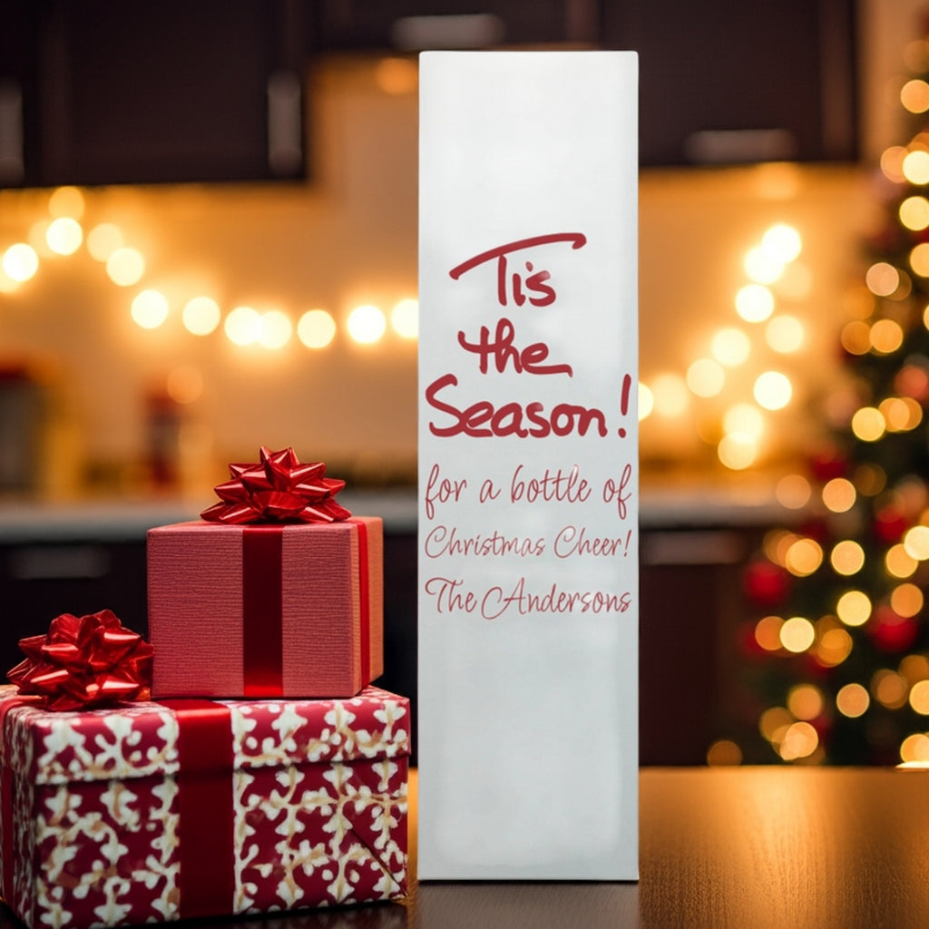 White wine bottle bag reading Tis the Season beside wrapped gifts and tree lights custom printed Christmas wine bottle gift boxes