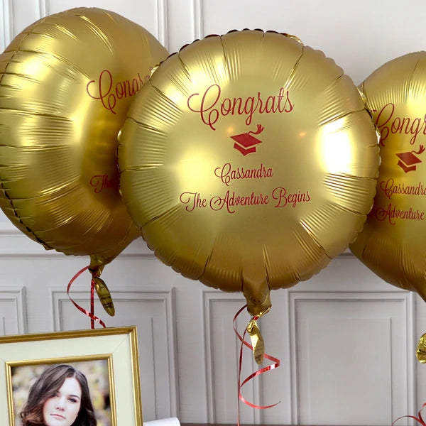 A vibrant graduation celebration image featuring three shiny gold balloons, each personalized with elegant red text that reads "Congrats" and "Cassandra The Adventure Begins," accompanied by a small graduation cap icon. The balloons are decorated with red and gold ribbons. In the foreground, a framed photo showcases the graduate herself. The backdrop includes a white paneled wall. Personalized Mylar balloons are ideal for indoor or outdoor graduation party decorations.