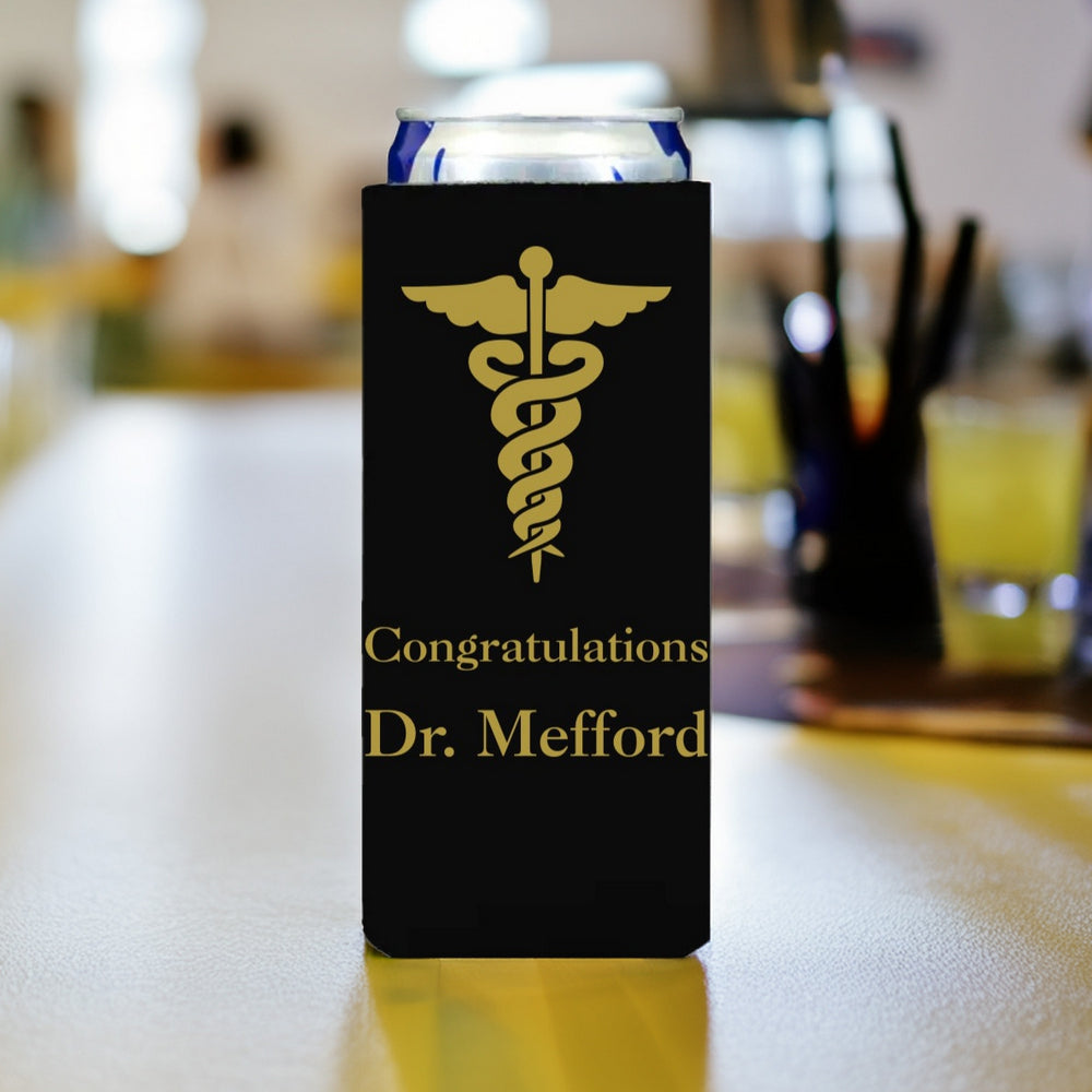 A slim can is nestled inside of a black slim can sleeve personalized for a medical school graduation celebration in gold print. The top of the sleeve is printed with a large caduceus medial symbol with "Congratulations Dr. Mefford" below in a bold italic font. The skinny can beverage is setting on a wood table at a banquet hall.