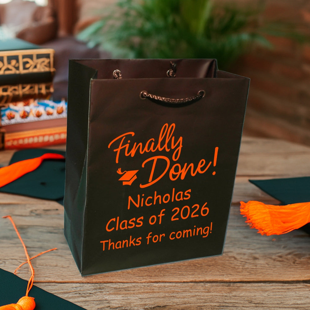 A brown Euro style gift bag personalized for a graduation party in orange print is setting on a dark wood table next to graduation caps with orange tassels. A stack of school books is visible in the background. A grad cap accents the large script words "Finally Done!" to exclaim the graduate's major achievement. Below the design is the graduate's name and wording "Class of 2026, Thank for coming!" to show the graduate's appreciation to their guests for coming to celebrate their milestone accomplishment.