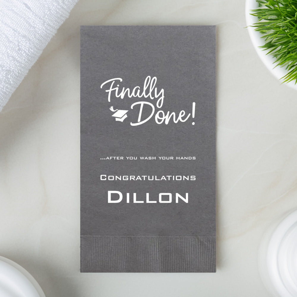 A pewter gray color paper hand towel rests on a marble vanity top in a bathroom for a graduation party. The guest towel is personalized with a calligraphy design with the offset wording "Finally Done!" accompanied by a small graduation cap. Three lines of text below the graduation design add a bit of fun to the graduation venue restroom with the wording "...After you wash your hands" and "Congratulations" above the graduate's name.