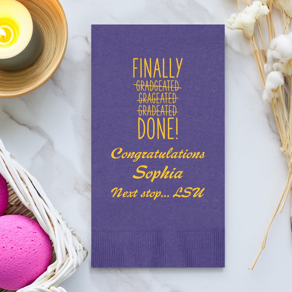 A purple paper guest towel playfully personalized in yellow print for a graduation is laying flat on a marble vanity top surrounded by a candle, hand soaps and dried flowers. The witty design on the napkin features the wording "Finally Done!" with the words "Gradgeated, Grageated, and Gradeated" crossed as they are spelled incorrectly. A fun idea for any student celebrating their graduation. Below the design are three lines of text congratulating the graduate with a "Next stop... LSU" message.