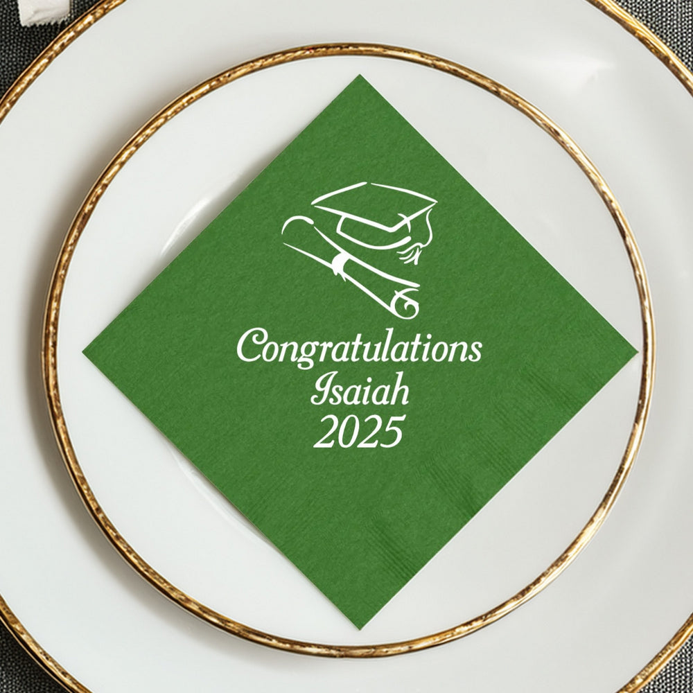 A festive green luncheon napkin oriented in a diamond shape personalized in white print for a graduation party. In the center of the napkin is a graduation cap and diploma graphic with the wording "Congratulations", the graduate's name and the graduation year "2025" in a a fun italic font. The napkin is laying on top of a white plate set with gold trim on a formal graduation party dining table.