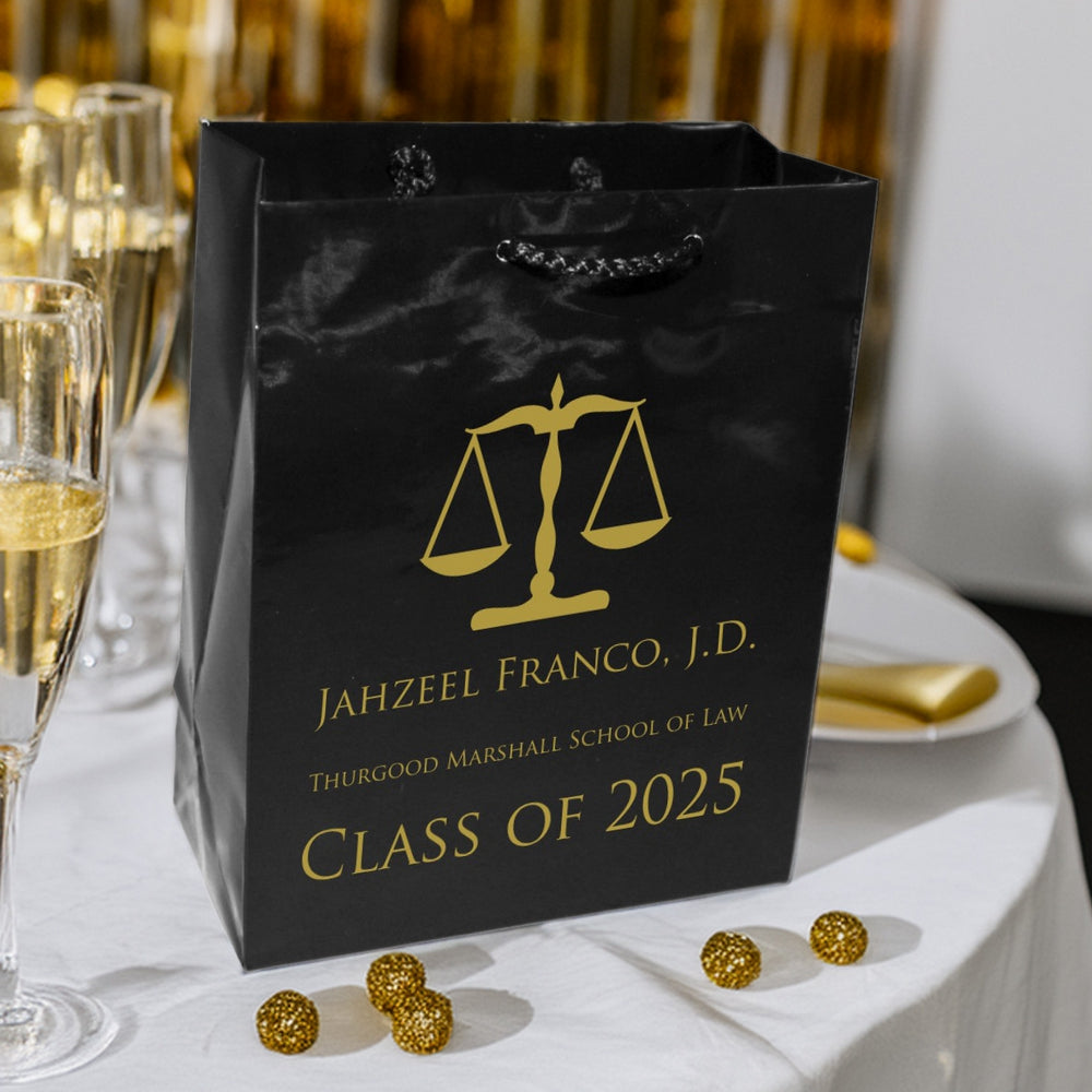 A glossy black Euro Tote gift bag with black braided handle personalized in gold print for law school graduation party guest gifts is setting next to glasses of champagne on a formal dinner table decorated with white linen tablecloth and gold napkins on white plates. A graphic of the scales of justice is prominently positioned above the graduate's name along with the law school name, and phrase "Class of 2025". The majestic looking bag conveys the law grad's major milestone accomplishment.