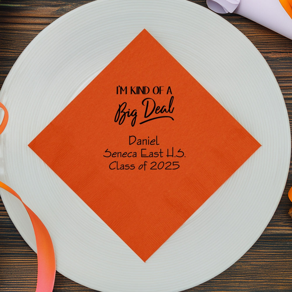 An orange paper dinner napkin custom printed for a graduation celebration is laying on a white plate that is resting on a dark, wood grain table decorated with orange ribbon. The napkin is personalized in black print with a fun graphic that displays the wording "I'm Kind Of A Big Deal" in a mix of italic and script fonts. Below the design in black print is three lines of custom text that includes the graduate's name, their high school name and "Class of 2025" in a fun italic font.