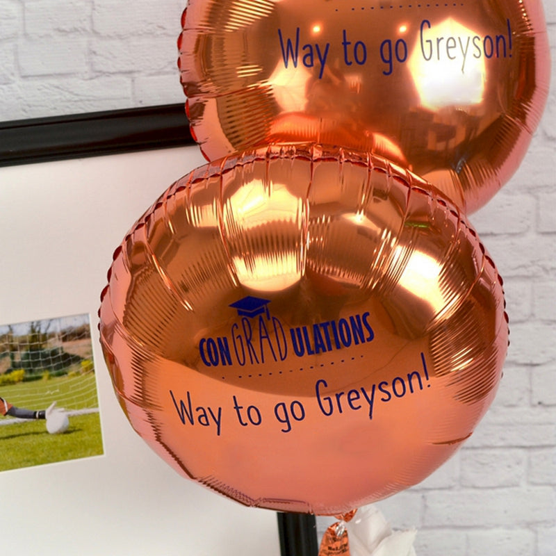 This charming image highlights an elegant graduation party decoration setup featuring two glossy rose-gold balloons, each inscribed with messages in blue text. The balloons proudly display "CONGRADULATIONS" for a witty play on words for a special graduation celebration, along with a small graduation cap icon, and the personalized phrase "Way to go Greyson!." Ideal for creating a memorable graduation party atmosphere, personalized balloons can be used to showcase your graduate's achievements.