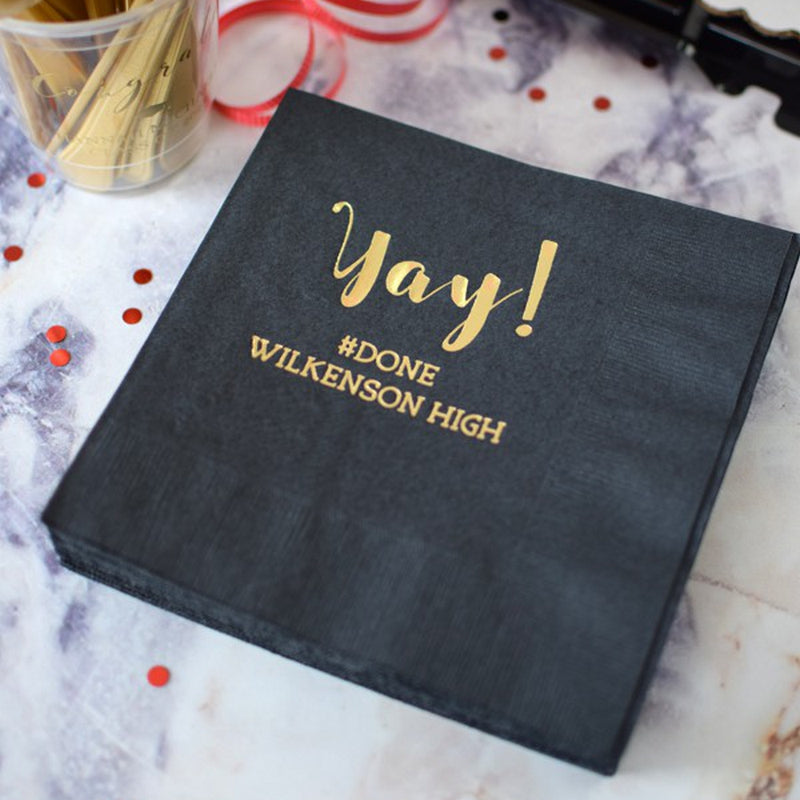 A black, square-oriented luncheon napkin personalized for a graduation party laying on a marble tabletop. Surrounding the napkin are grad party decorations including a personalized clear plastic cup filled with drink stirrers, red ribbon and confetti. The napkin is customized with a "Yay!" script graphic and two lines of custom text below with "hashtag DONE" and high school name in gold print.