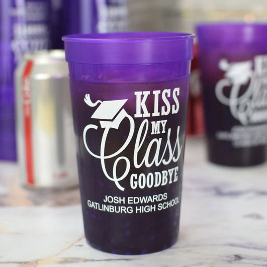 Translucent purple color 22 oz. graduation party cups personalized with kiss my class goodbye design and 2 lines of custom text in white print on table with drinks