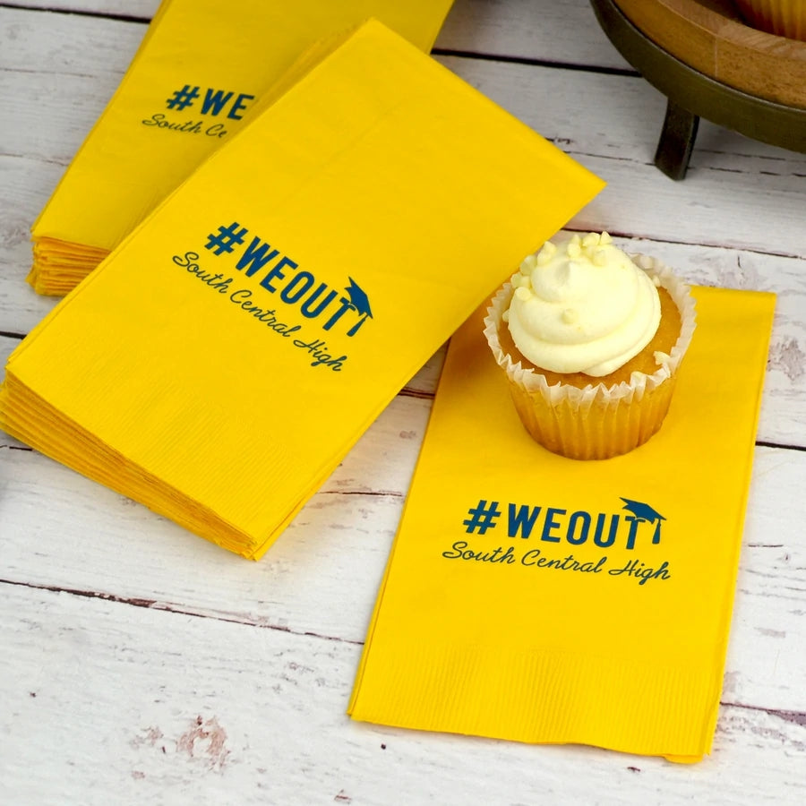Yellow color paper guest towel napkins personalized for a graduation party laying on a white wood table with a cupcake setting on one of the napkins. The guest towels are customized in hunter green print with a hashtag we out design featuring a graduation cap above the letter "T". Below the hashtag design is a custom line of text displaying the graduate's high school name.