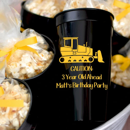 Black 32 oz jumbo stadium kids birthday cups personalized with bulldozer design and 3 lines of text in yellow print filled with popcorn favors