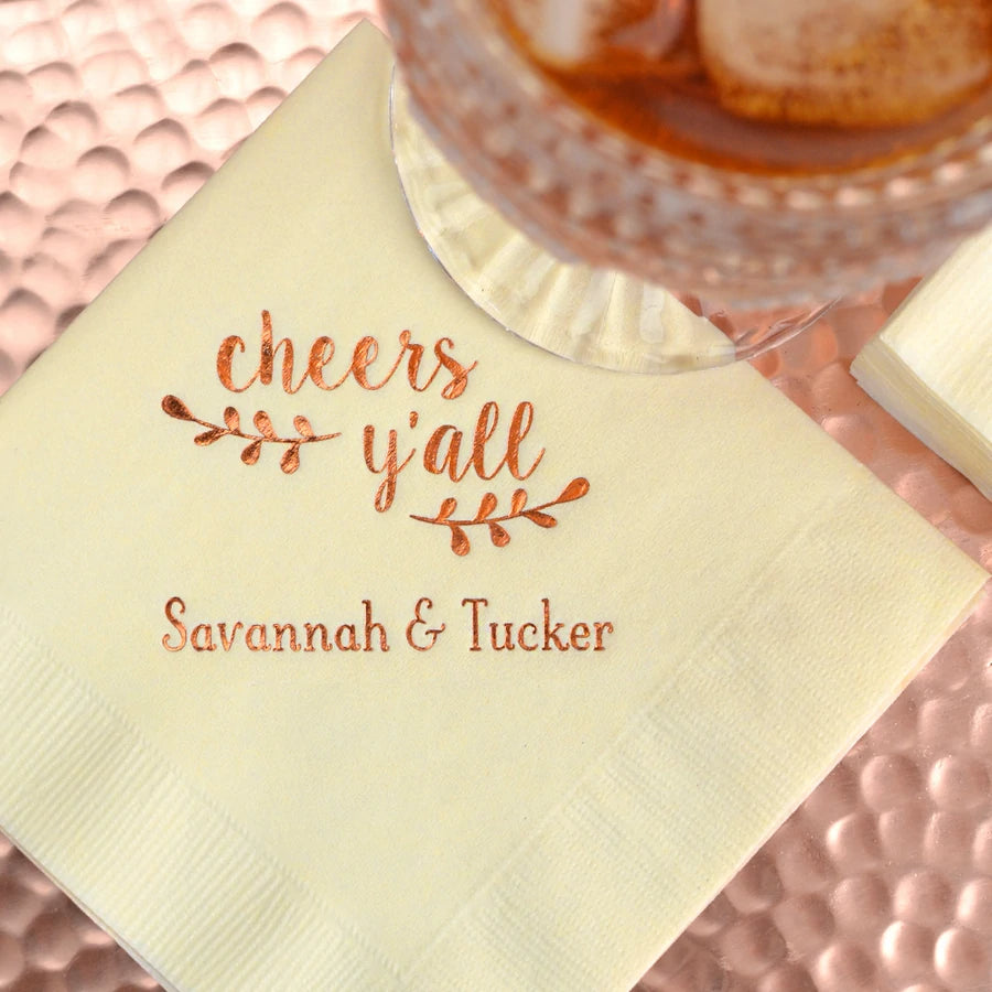 beverage sitting on ivory wedding cocktail napkin personalized with cheers yall design and bride and groom name in copper print