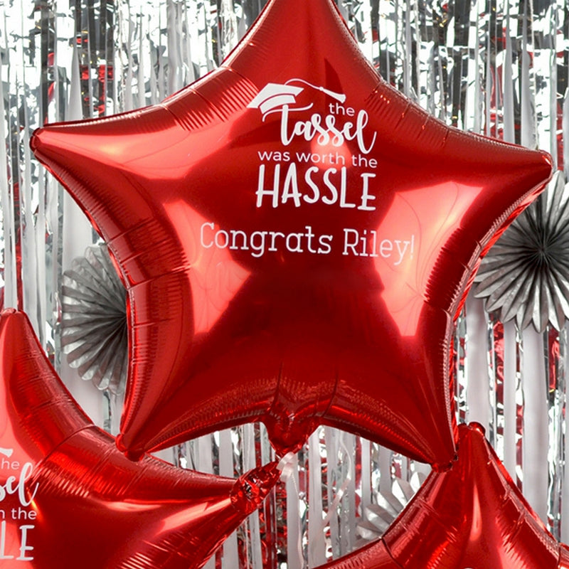 Metallic red star shape Mylar balloons personalized for a graduation party in white print with "the Tassel was worth the Hassle" graduation design and custom text "Congrats [Graduate's Name." The balloons are floating in front of a silver streamers background.