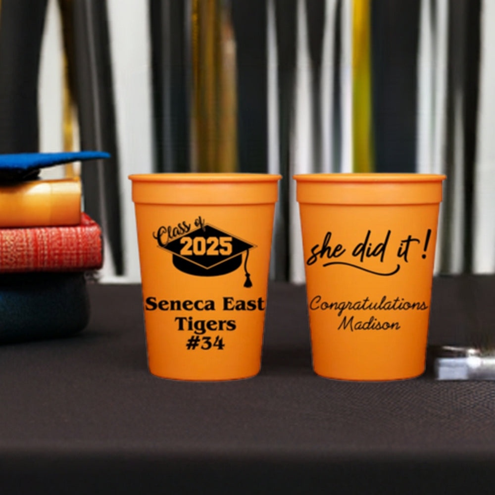 Two small vibrant orange cups personalized for serving grad party drinks are set on a black tabletop with a blue graduation cap set on a stack of books, with gold and black streamers in the background. The cup on the left proudly displays "Class of 2025" with the school name, mascot, and a sporty "#34," while the cup on the right celebrates the special grad with a fun "SHE DID IT!" design followed by custom text that reads "Congratulations Madison" in "Playful" font type.