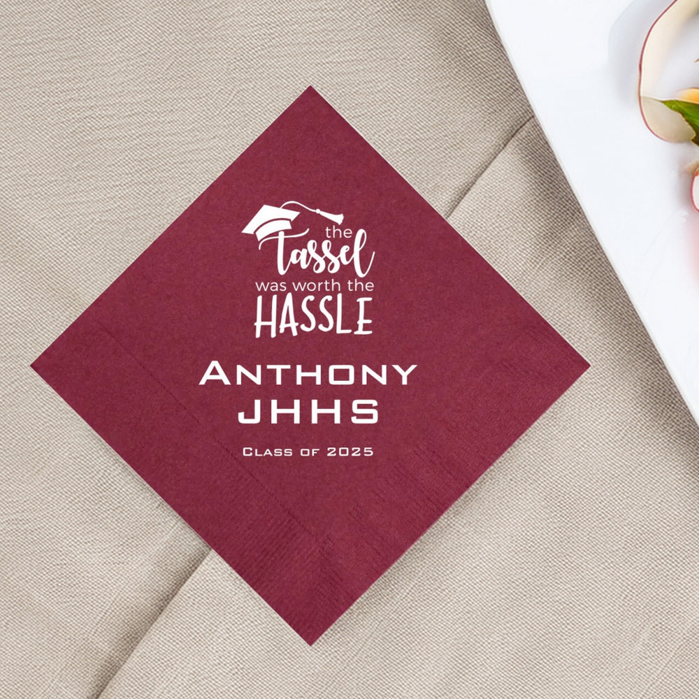 A burgundy color paper luncheon napkins personalized for a graduation party in white print. A graphic design at the top features the working "the Tassel was worth the HASSLE." Three lines of custom text below the graphic include the graduate's name, high school initials and "Class of 2025" in a bold, block font. The burgundy and white colors give the napkins a classic and elegant look. The napkin is laying on a taupe linen tablecloth next to a plate.