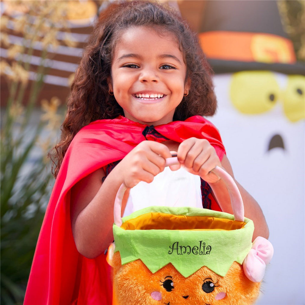 Smiling child in red cape holding personalized trick or treat candy bags shaped like a pumpkin with the name Amelia