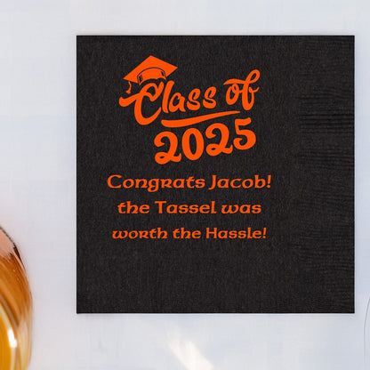 A black beverage napkins personalized for a graduation party in orange print in square orientation. A graduation graphic "Class of 2025" is printed above three lines of custom text. The first line reads "Congrats [graduate name]. The second line of text reads "the Tassel was". The third line reads "worth the Hassle!" The font used for the three lines of custom text is a modern block font. The napkin is laying on a white linen tablecloth next to a glass holding a beverage.