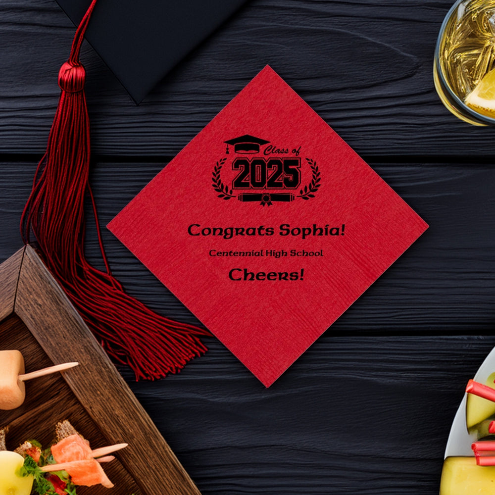 A red paper cocktail napkin personalized for a graduation party. The napkin is customized in black print with "Class of 2025" graphic art featuring a graduation cap and diploma with laurel sprigs on the left and right of the design. Below the design are three custom lines of text with congratulations to the graduate, high school name and "Cheers!" in a modern semi-script font. The napkin is on a black table with drinks, hors d'oeuvres, and a crimson red tassel extending from a black graduation cap.