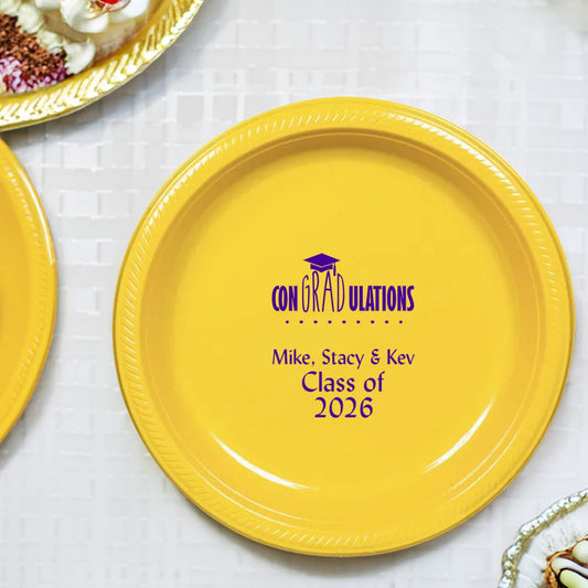 A yellow plastic dessert plate personalized for a 2026 graduation party features a design with the word "ConGRADulations" for a fun play on the word "congratulations" for the graduate. A grad cap graphic rests atop "GRAD" in the word "Congradulations" with a series of ornamental dots below. Below the design are three lines of text including three graduates' names, and "Class of 2026" in a fun, simple italic font.