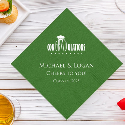 A festive green color beverage napkin personalized for a graduation party is laying on a wide wood table next to drinks and food. The napkin is customized in white print with a graphic design that features the working "conGRADulations" with multiple dots underneath. A graduation cap is suspended over "GRAD". Below the graduation design is three lines of custom text with the working "[graduates' names], CHEERS TO YOU! CLASS OF 2025.