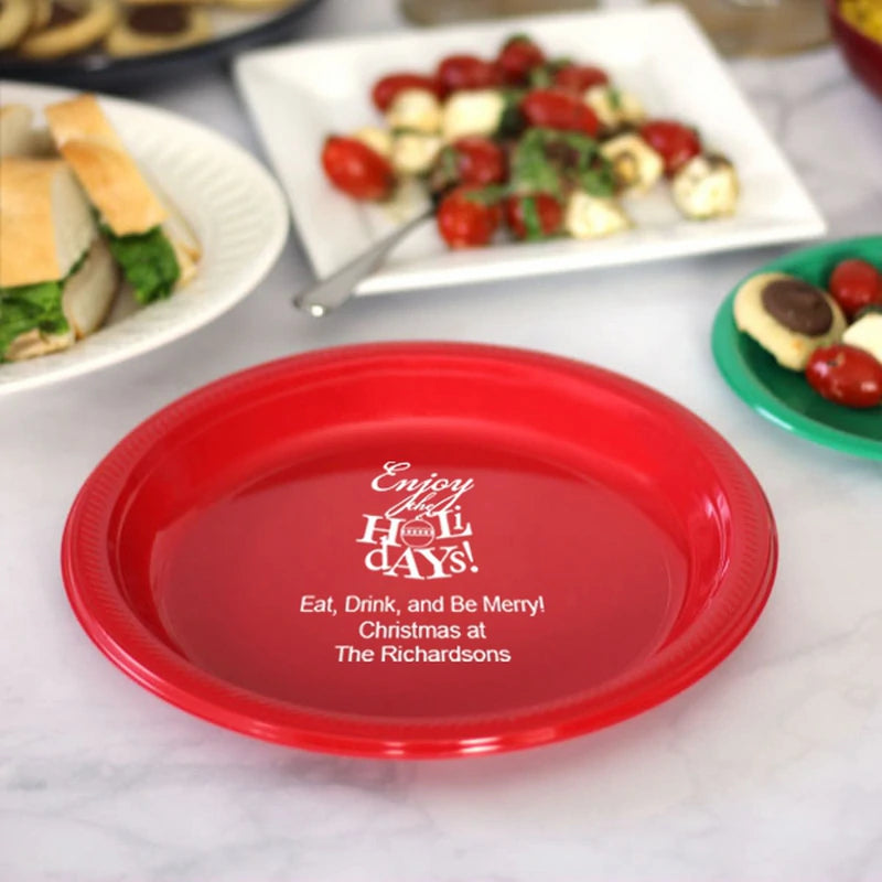 Red disposable plastic plate personalized for Christmas with Enjoy the Holidays design and cusotm message in white print on a party table with assorted appetizers and cookies in the background.