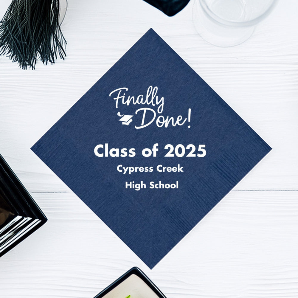 A blue cocktail napkin personalized for a graduation party in white print. A script design graphic with the wording "Finally Done!" features a small graduation cap amidst the offset wording. Below the design are three lines of custom text with the wording "Class of 2025, Cypress Creek, High School. The napkin is laying on a white wood tabletop with a glass, graduation cap tassel, and plates.