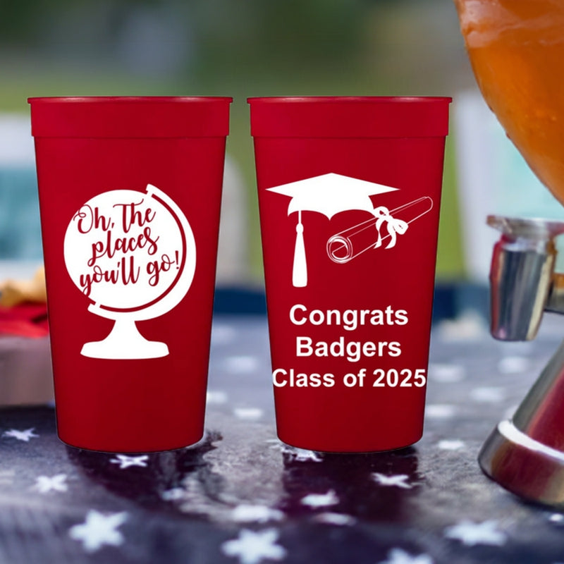 Two tall red color plastic stadium cups customized for a graduation party. The cup on the left displays a globe graphic with the words "Oh, the places you'll go!" in white print. The cup on the left side features a graduation cap and diploma design. Below the design is the text "Congrats Badgers Class of 2025" in bold block white lettering. the cups are setting next to a drink dispenser on an outdoor table covered with a blue tablecloth with white stars for a backyard graduation party.