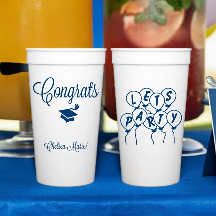 Two large custom printed white plastic tumbler cups in front of drink dispensers on a table at an outdoor graduation party. The cup on the left is personalized with the Word "Congrats" above a graduation cap and the graduate's name in script lettering in navy print. The cup on the right is personalized with a party balloons graphic with letters in the center of each balloon spelling "LETS PARTY" in navy print.