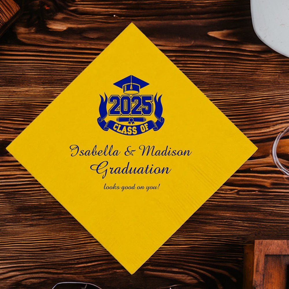 A yellow beverage napkin personalized for a 2025 graduation in blue print. The graduation design features a grad cap above block numbers "2025". Below the block numbers is a diploma. The graphic includes a ribbon that wraps around the diploma and "2025" with the working "CLASS OF". Below the graduation "Class of" design is three lines of custom text that includes the wording "graduates' names, Graduation looks good on you!" The napkin is laying on a dark wood picnic table set up for a backyard grad party.