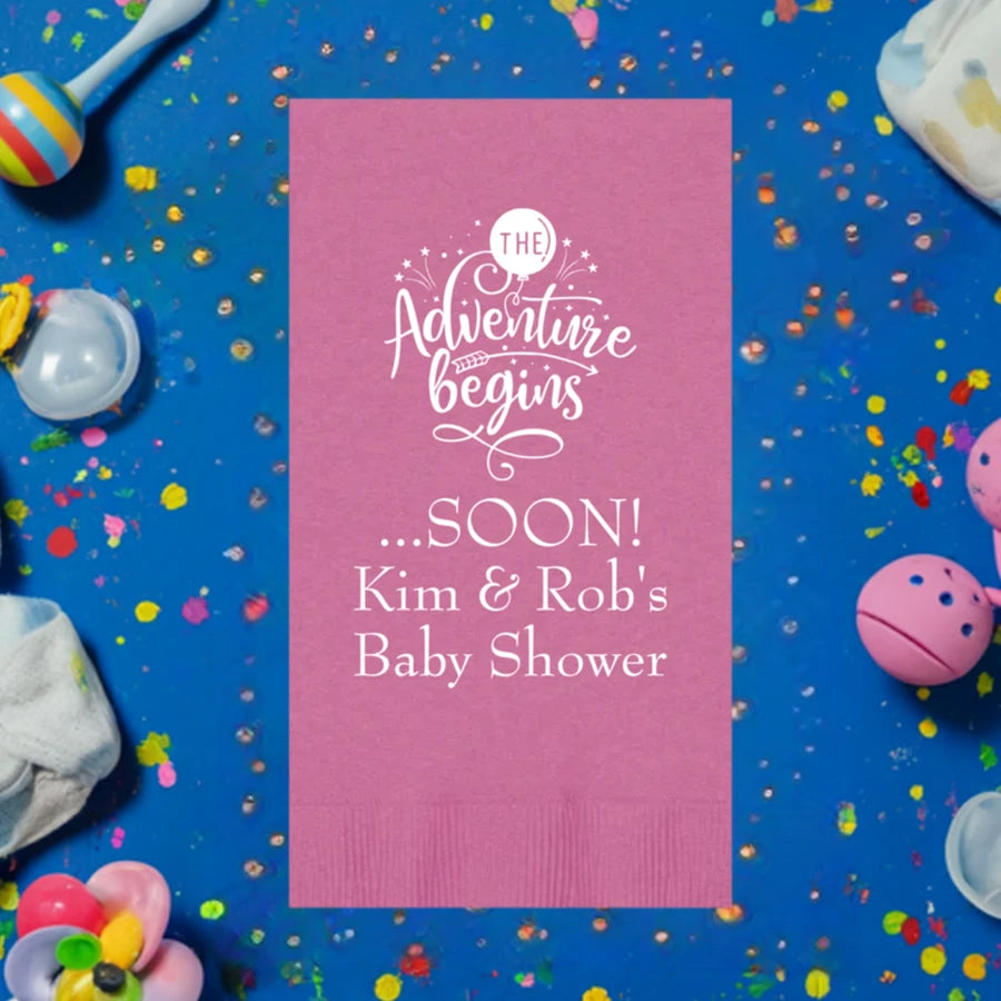 Bright pink color paper baby shower guest towel personalized with the adventure begins design and 3 lines of text in white print