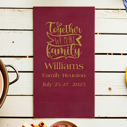 A burgundy color paper family reunion guest towel napkin personalized with together we make family design and 3 lines of custom text in gold print on a picnic table