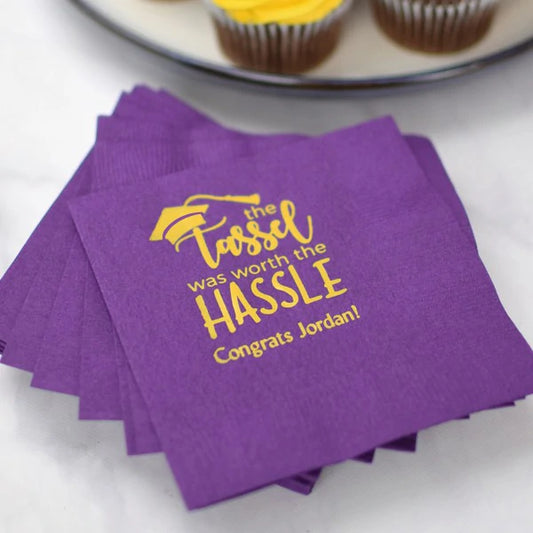 A stack of purple color beverage napkins custom printed in yellow ink for a graduation celebration. A design "the Tassel was worth the HASSLE" and "Congrats [graduates name] is displayed on the napkin in square print layout. The napkins are setting on a table with white linen tablecloth below a plate of cupcakes.