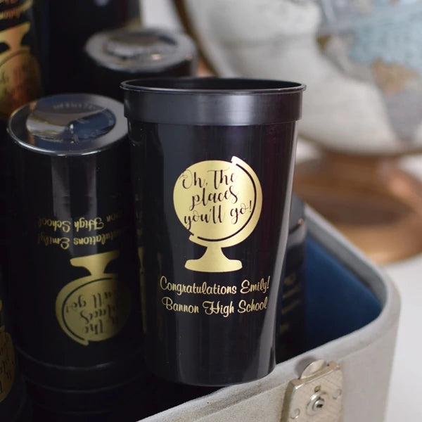 Black color 22 ounce graduation stadium cups personalized with oh the places you will go design and 2 lines of custom text in gold print stacked in suitcase