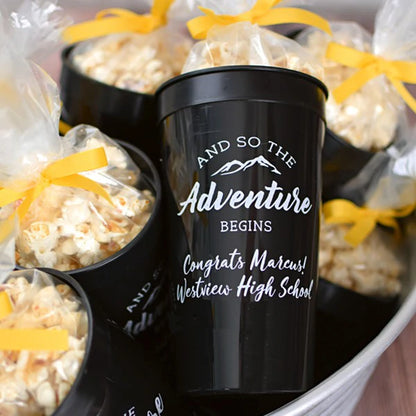 Black 32 ounce stadium graduation favor cups personalized with adventure begins design and 2 lines of custom text in white print filled with popcorn