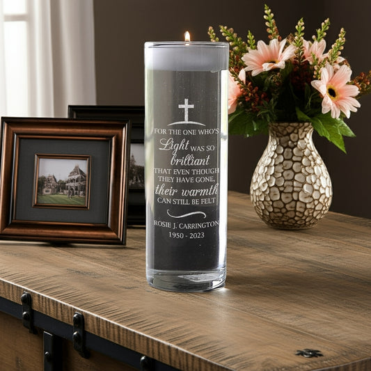 Personalized memorial candle vase with engraved tribute and cross on a wooden table next to photo frames and flower vase