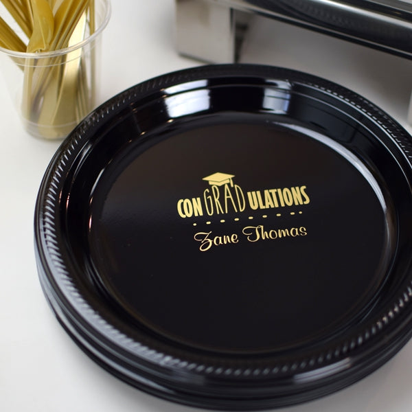 This sleek black plastic graduation dinner plate is personalized with a golden message for the graduation celebrant: "CONGRATULATIONS [Graduate Name]!" A matching gold graduation cap adds a celebratory flair. Set beside golden utensils, this graduation keepsake plate marks a triumphant milestone, ready to be filled with delicious memories as the graduate steps into a bright future!